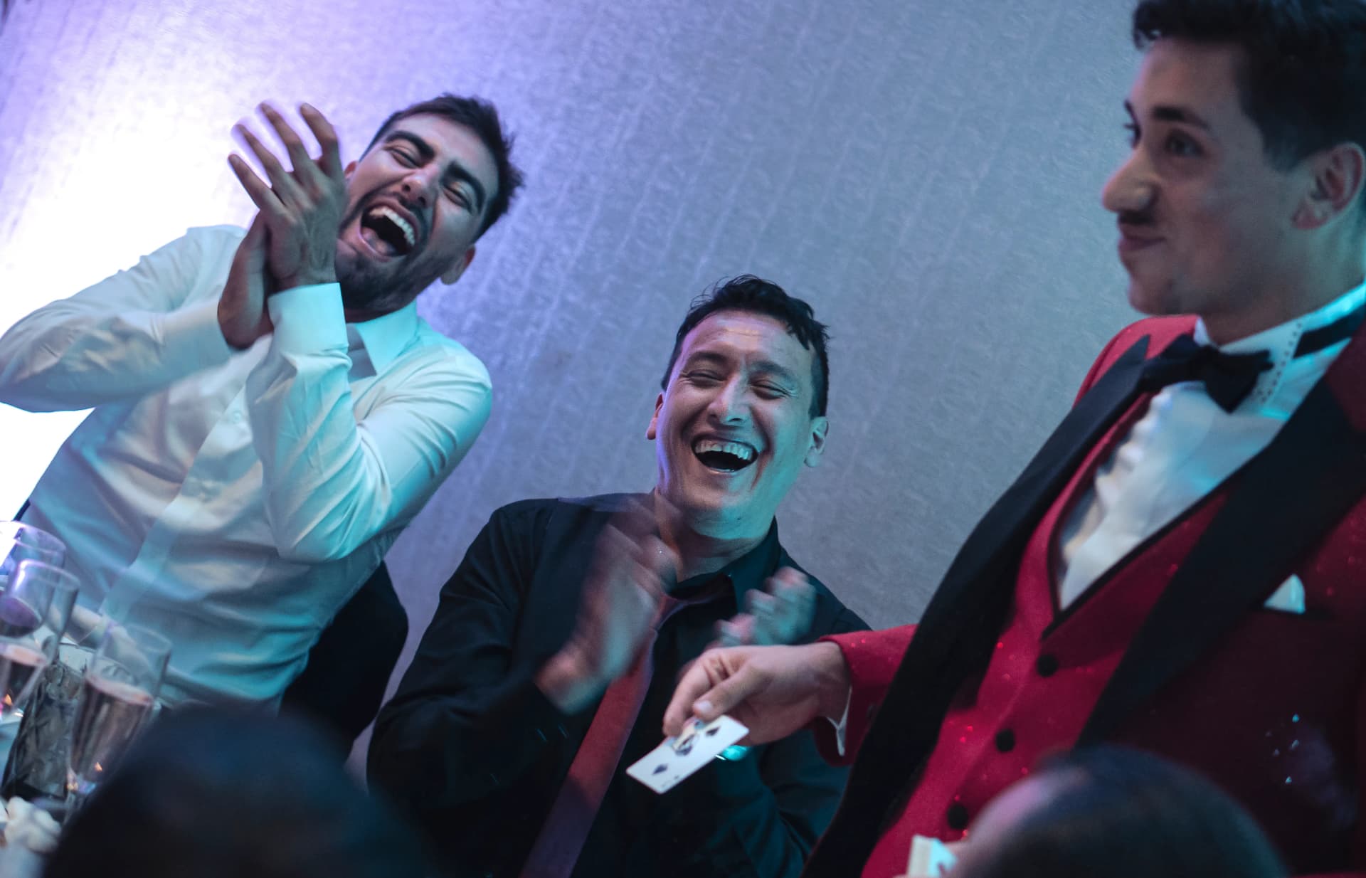 Dos invitados riendo en primer plano junto a Aníbal con traje rojo